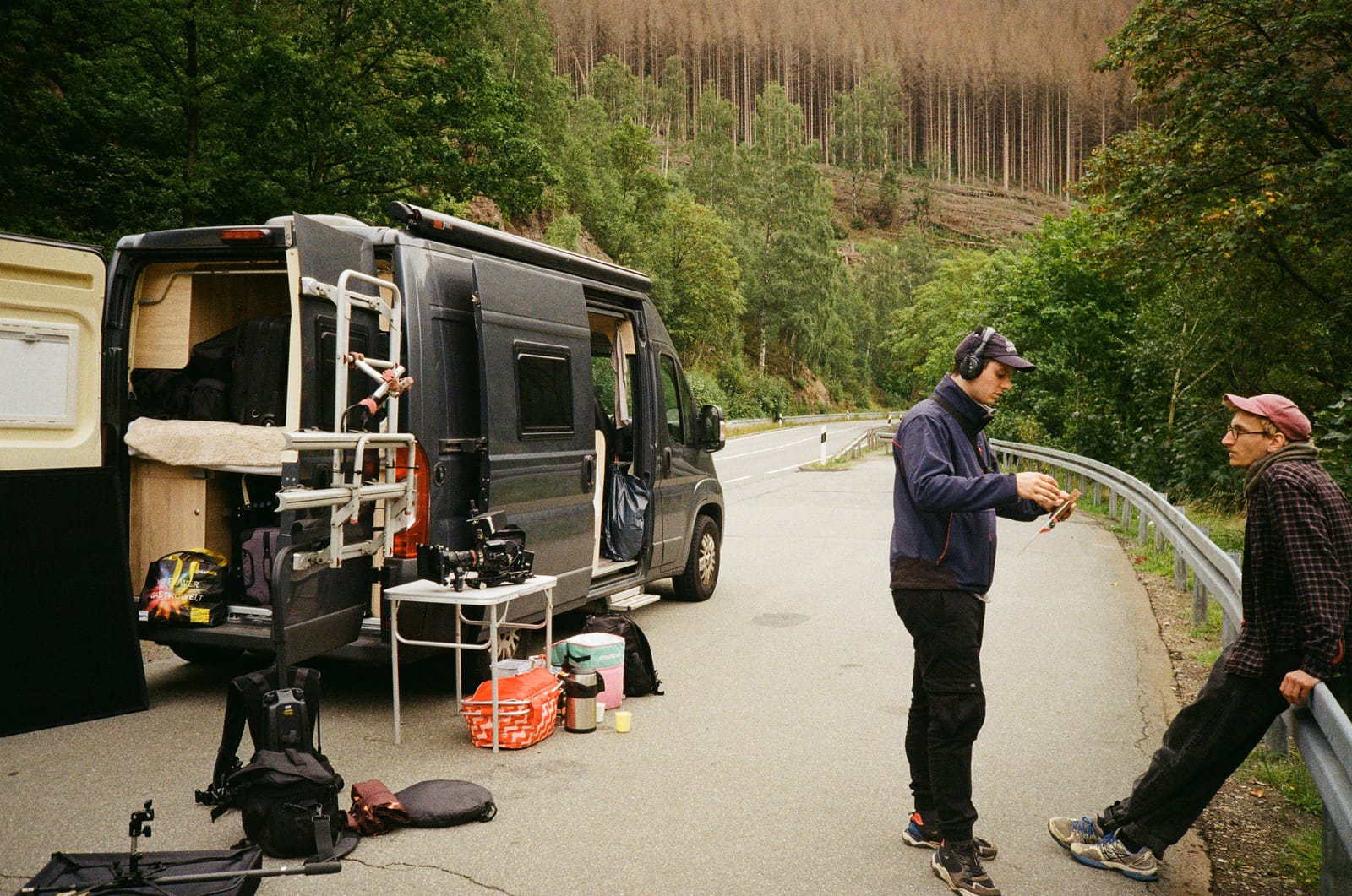 Crew checking sound by an open camper van, camera rig set up on a folding table at a forest roadside