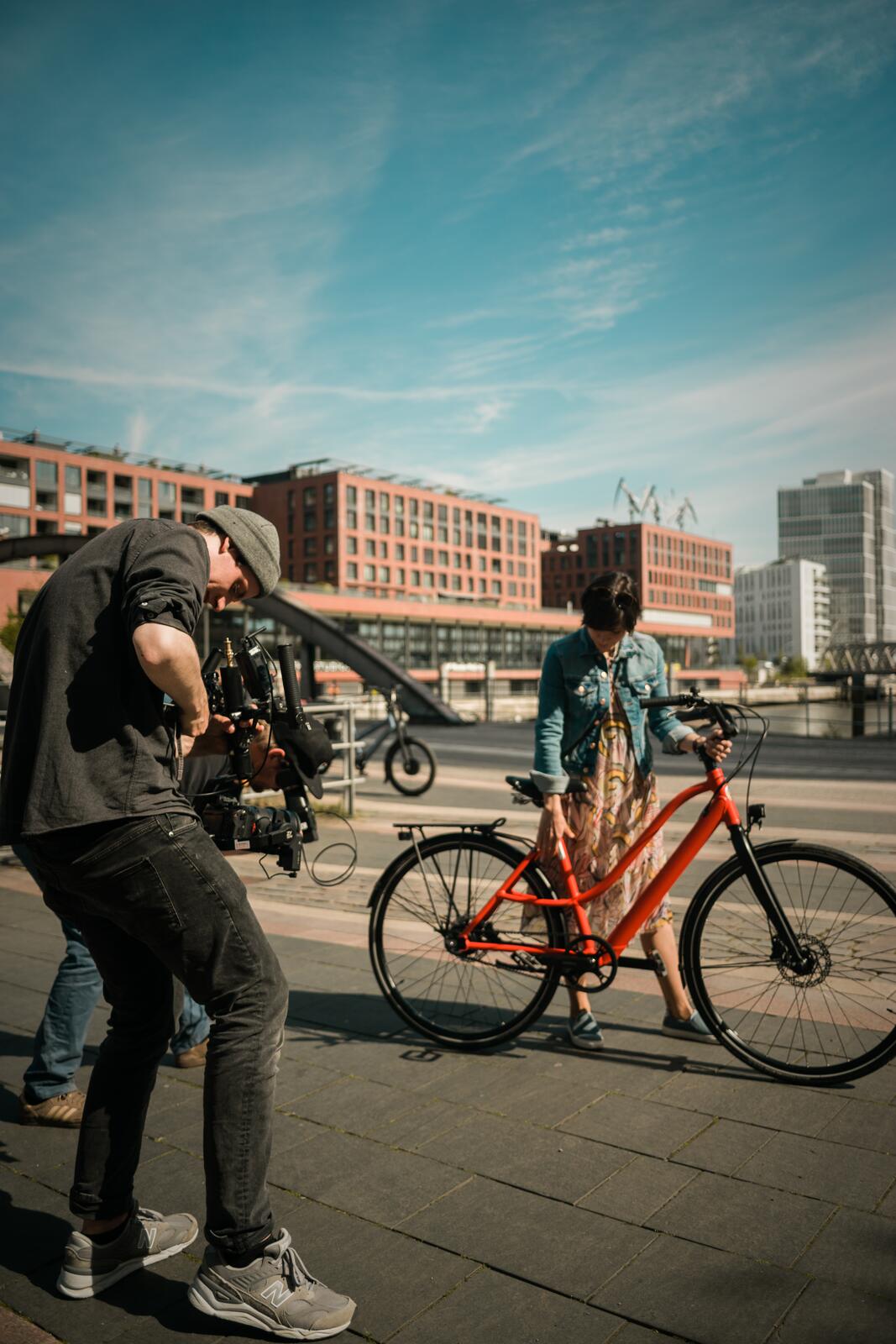 Operator filming low on a gimbal as a woman walks a red bicycle along a harbour promenade