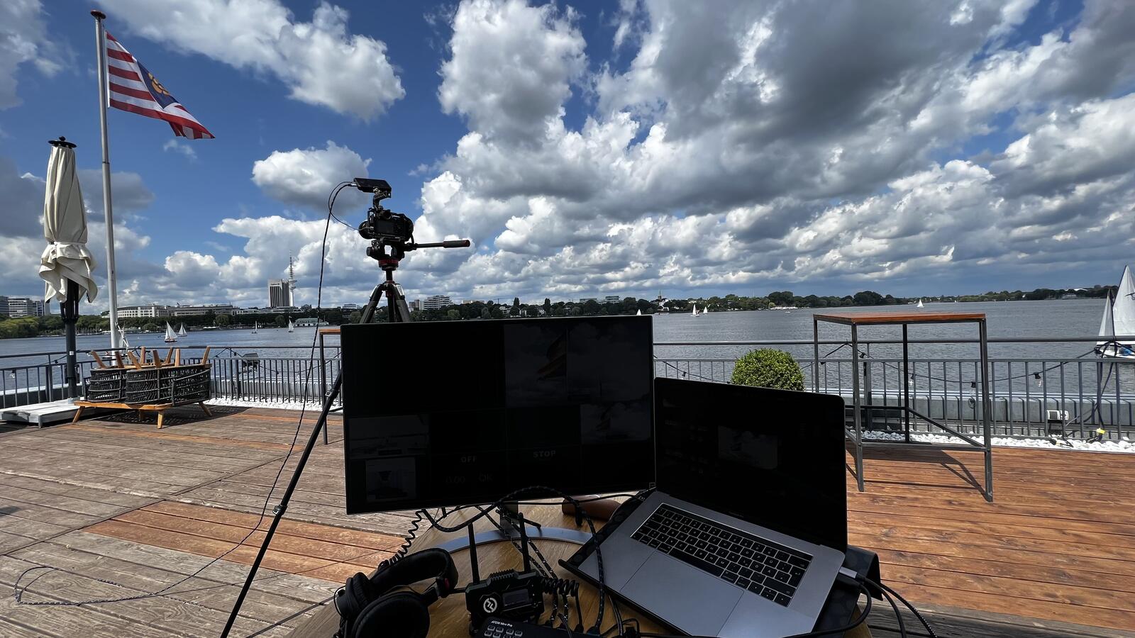 Video village on a lakeside jetty: monitor and laptop on a table, a camera on a tripod, big clouds overhead