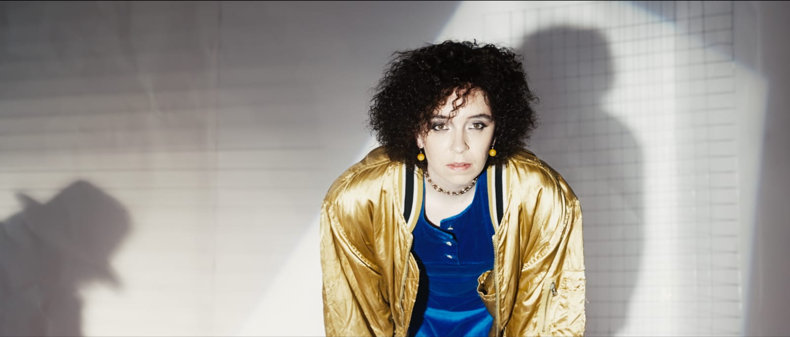 Portrait of a woman in a gold jacket against a white tiled wall